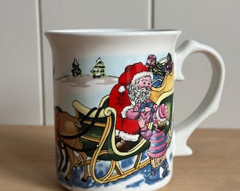 May include: A white ceramic mug featuring a colorful illustration of Santa Claus in a green and yellow sleigh pulled by a reindeer. Santa is handing a gift to a child wearing a striped sweater and blue boots in a snowy landscape with pine trees.