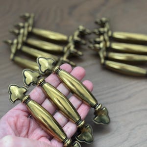 May include: Several antique brass cabinet pulls with a decorative cloverleaf design. The pulls have a curved, cylindrical handle and are arranged on a wooden surface. The brass finish gives them a vintage look, suitable for furniture restoration or adding a classic touch.