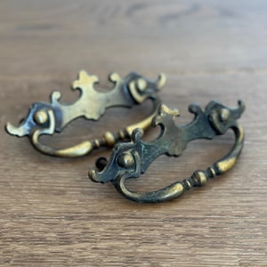May include: Two antique brass drawer pulls with ornate, curved designs. The pulls have a dark patina with hints of green and gold, and are set against a wooden surface. These vintage hardware pieces are ideal for furniture restoration.