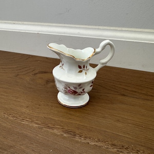 May include: A small, white ceramic creamer with a scalloped rim and a handle. The creamer features a floral design in shades of red and brown, with gold trim. The creamer is approximately 4 inches tall.