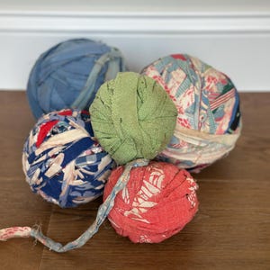 May include: Five balls of fabric strips in various colors and patterns. One ball is solid sage green, one is solid blue, one is solid red, one features a blue and white floral print, and one has a multi-colored pattern with red and blue accents.