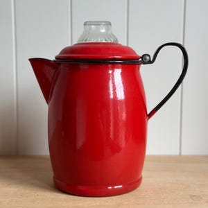 May include: A bright red enamel coffee percolator featuring a black rim and a matching black curved handle. The pot includes a clear glass knob on the lid, a side spout, and a smooth, glossy finish throughout the exterior of the metal body.