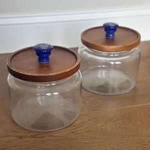 May include: Two clear glass jars with wooden lids. Each lid has a blue knob with a white floral design. The jars sit on a wooden surface, and the background is a white wall.