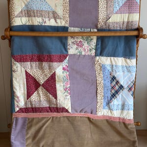 May include: A patchwork quilt featuring a variety of fabric squares in shades of blue, lavender, burgundy, and cream. The design includes floral patterns, plaid, and solid colors, with a horizontal pink velvet ribbon accent and a brown fabric border.