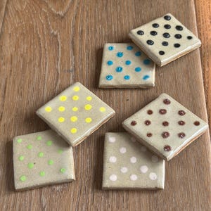 May include: Five square ceramic tiles with a glossy beige finish, each featuring a distinct pattern of polka dots in different colors including black, blue, yellow, green, brown, and white, arranged in a grid-like formation on each individual tile.