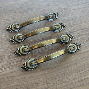 May include: Four antique brass drawer pulls with a curved handle design. Each pull features decorative end pieces and a rectangular center. The hardware is arranged on a wooden surface.