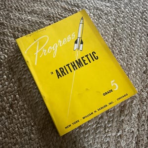 May include: A vintage yellow textbook titled "Progress in Arithmetic" for Grade 5. The cover features a rocket illustration and the publisher's information. The book is lying on a textured, neutral-colored surface.