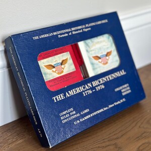 May include: A blue box set of The American Bicentennial 1776-1976 playing cards by U.S. Games Systems, Inc. The box features a cutout window displaying two decks of cards with eagle and shield illustrations, and includes text about historical figures.