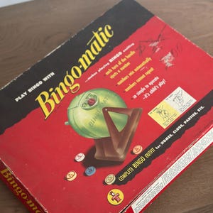 May include: A vintage Bingo-matic game set in a red box with yellow and black text. The game includes a green ball dispenser and brown number selector. The box reads "COMPLETE BINGO OUTFIT for HOMES, CLUBS, PARTIES, ETC."
