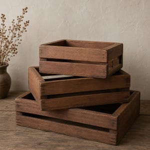 May include: Three wooden crates stacked on top of each other. The crates are rectangular with slatted sides and a dark brown finish. The crates are empty and appear to be handmade. The background is a neutral color.