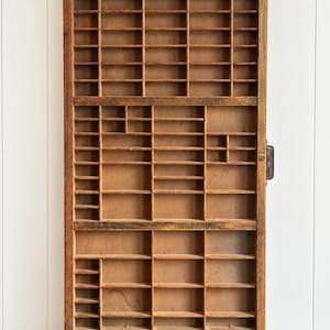 May include: A rectangular wooden printer's tray featuring a grid of numerous small, open compartments of varying sizes. The wood has a natural brown finish with visible grain and a weathered texture throughout the entire structure.