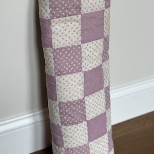 May include: A rectangular quilted fabric piece featuring a checkerboard pattern of solid dusty mauve squares alternating with cream-colored squares that have a small, repeating floral print. The fabric has visible stitching lines throughout.