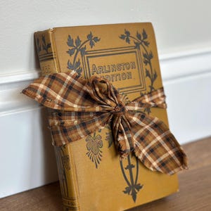 May include: A vintage mustard-yellow hardcover book titled Arlington Edition, featuring black floral and geometric decorative patterns. The book is wrapped horizontally with a brown, black, and white plaid fabric ribbon tied in a bow.