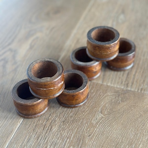 May include: Six wooden napkin rings in a warm brown hue are arranged on a light-colored wooden surface. Each ring is cylindrical with a visible grain and a lighter-colored band around the middle, suggesting a natural, rustic aesthetic.