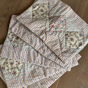 May include: A stack of quilted square placemats. Each placemat features a patchwork design with floral and striped patterns in cream, pink, and blue. The placemats are arranged on a wooden surface, showcasing their layered construction.