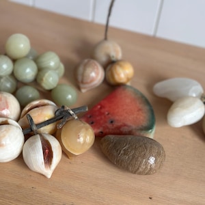 May include: A collection of decorative stone fruit sculptures including a cluster of pale green grapes, several carved stone peaches with visible seams, a polished watermelon slice, a brown textured pear, and a cluster of smooth white stone fruit.