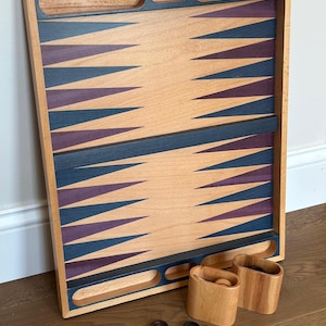 May include: A wooden backgammon board with a light wood finish, featuring blue and purple triangular points. Includes wooden cups and checkers in light and dark wood tones. The board has a rectangular shape with cut-out handles.