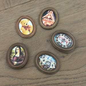 May include: Five small, oval-shaped miniature framed pictures. The frames are brown. The pictures include a teddy bear, a painting of a woman, and other colorful images. The miniatures are arranged on a wooden surface.