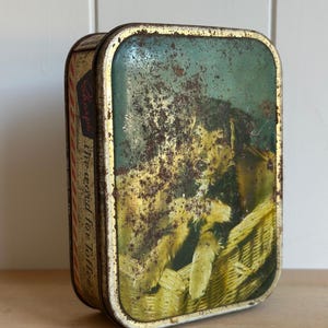 May include: A rectangular metal tin with rounded corners featuring a weathered green and yellow illustration of a dog in a basket. The side of the tin displays the text Sharps the word for Toffee with red and white diagonal stripes.