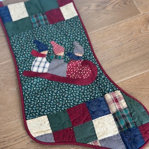 May include: A quilted Christmas stocking featuring a patchwork border of squares in red, green, blue, and cream fabrics. The center panel is dark green with a small floral print and an appliqué of three figures riding a red sled together.