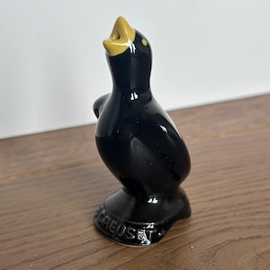 May include: A glossy black ceramic bird figurine with a yellow beak and accents. The bird is standing on a base with the word "CREUSET" embossed on it. The figurine has a smooth, reflective surface.