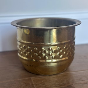 May include: A polished brass planter with a wide, flat rim and a rounded body. The middle section of the exterior features a textured band of raised, evenly spaced circular bumps, while the top and bottom sections remain smooth and reflective.