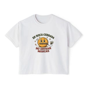 May include: White t-shirt with a graphic featuring a yellow smiley face with a stitched mouth, surrounded by stars and a fly. The text reads "EN BOCA CERRADA NO ENTRAN MOSCAS" in a vintage style.