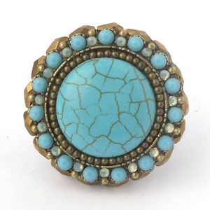 以下が含まれることがあります： 大きなひび割れたターコイズストーンを中央に配した、丸いアンティークスタイルのリング。リングは、ブロンズ色の金属フレームにセットされた、小さな水色と虹色のビーズで囲まれています。リングは、詳細でテクスチャード加工されたデザインです。