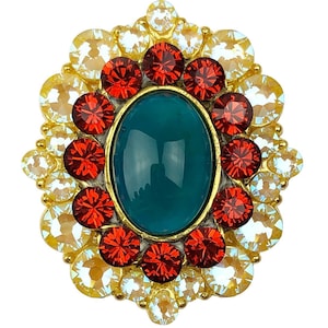 Może przedstawiać: Złota broszka z turkusowym owalnym kaboszonem otoczonym czerwonymi i przezroczystymi cyrkoniami.