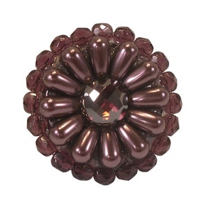 以下が含まれることがあります： 中央に濃い紫色のクリスタルがついた、丸い紫色の花の形をしたブローチ。ブローチは小さな丸いビーズで作られており、光沢のある仕上げになっています。