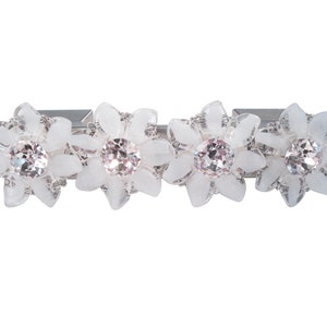 Puede incluir: Una pinza para el pelo plateada con tres adornos en forma de flor blanca, cada uno con un strass transparente en el centro.