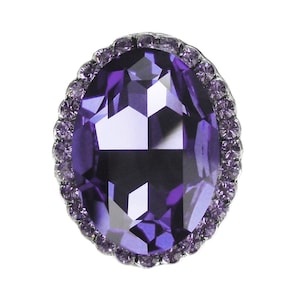 Puede incluir: Una gran piedra preciosa ovalada de color púrpura con un diseño facetado. La piedra preciosa está rodeada por un borde de piedras preciosas púrpuras más pequeñas.
