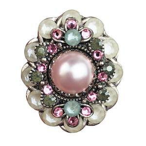 Puede incluir: Broche plateado con una perla rosa central, rodeada de gemas rosas, verdes y claras. El broche tiene un diseño floral con un borde festoneado, creando una estética vintage o antigua. Las gemas son redondas y están muy juntas.