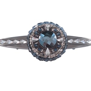 Op de afbeelding: Zilverkleurige haarspeld met een centrale, facetgeslepen, teal-kleurige edelsteen, omringd door kleinere blauwe en heldere kristallen. De haarspeld heeft een decoratief, cirkelvormig middelpunt en is versierd met druppelvormige kristallen aan de zijkanten.