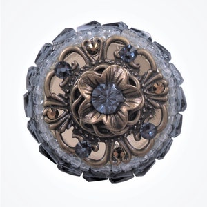 May include: A round, decorative button with a silver and gold floral design. The center of the button features a large, dark blue rhinestone surrounded by smaller rhinestones. The button is surrounded by a ring of clear, faceted beads.