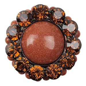 Könnte beinhalten: Ein runder, brauner Edelstein-Cabochon, umgeben von einem Ring aus kleinen, facettierten, bernsteinfarbenen Strasssteinen.