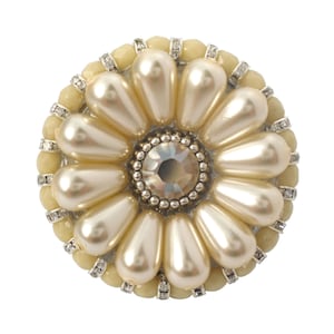 以下が含まれることがあります： クリーム色の花の形をしたブローチ。大きなクリスタルの中心にフェイクパールが並んでいます。ブローチは金属製で、光沢のある仕上げになっています。