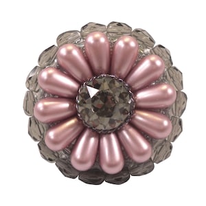 以下が含まれることがあります： 中央に大きな濃いグレーのラインストーンがついた、ピンクの花の形をしたブローチ。ブローチは、小さなクリアなラインストーンの輪で囲まれています。