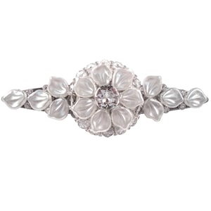Könnte beinhalten: Eine silberne Haarklammer mit einem weißen Blumendesign. Die Blume hat eine klare Strasssteine ​​in der Mitte und ist von kleineren weißen Blütenblättern umgeben.