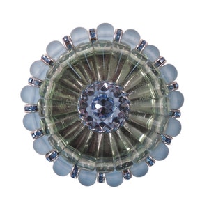 Könnte beinhalten: Ein runder, blauer Glasknopf mit einem großen, klaren Kristall in der Mitte. Der Knopf ist von einem Ring aus kleineren, blauen Kristallen umgeben.