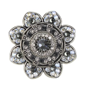 Puede incluir: Un broche de plata en forma de flor con un cristal central rodeado de cristales más pequeños. El broche tiene un diseño detallado con un patrón negro y plateado.