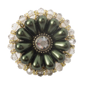 Peut inclure: Une broche en forme de fleur verte avec un grand cristal au centre, entouré de strass transparents.