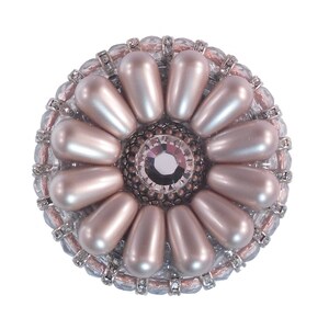 Puede incluir: Un broche redondo de tono plateado con un cristal central rodeado por un anillo de perlas falsas. Las perlas están dispuestas en forma de flor.
