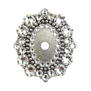Puede incluir: Adorno decorativo ovalado plateado con un agujero central. El borde está adornado con un anillo de pequeñas cuentas redondas y un anillo exterior de cristales grandes, transparentes y facetados. El diseño es simétrico y refleja la luz.