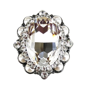 以下が含まれることがあります： 小さなラインストーンと白い真珠の輪で囲まれた、大きな透明なクリスタルラインストーン。ラインストーンと真珠は銀色の金属にセットされています。