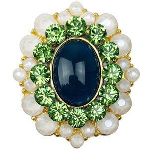 Peut inclure: Une broche dorée avec une grande pierre ovale bleu foncé entourée de petites pierres vertes et de perles blanches.