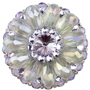 以下が含まれることがあります： 丸い装飾的なブローチ、花の模様。ブローチは、中央にファセット加工された薄紫色のクリスタル、透明な虹色のクリスタル、小さな紫色のクリスタルで囲まれています。ブローチは白い背景に配置されています。