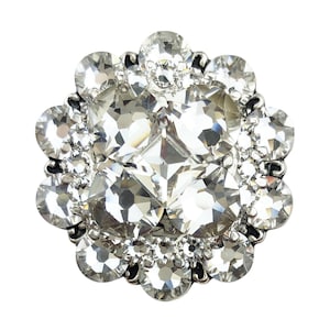 Puede incluir: Un botón redondo y brillante, de color plateado, con un diseño floral. El botón presenta un gran cristal central facetado rodeado de cristales transparentes más pequeños, creando un efecto deslumbrante. Ideal para embellecer ropa o accesorios.