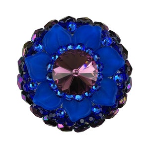 Peut inclure: Une broche en forme de fleur bleue et violette avec un grand cristal violet facetté au centre. La broche est en verre et en strass.
