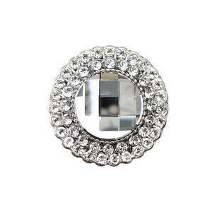 Könnte beinhalten: Ein runder, silberfarbener, dekorativer Knopf mit einem klaren, facettierten Kristall in der Mitte, umgeben von einem Ring aus kleineren Kristallen.